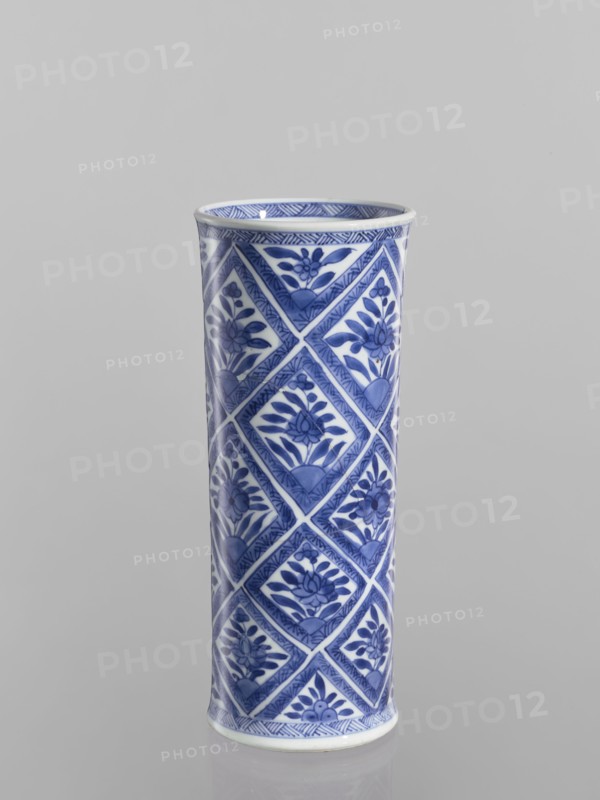 Vase, Chinese, blue-and-white porcelain, ceramics; cylindrical vessel with geometric and floral ornaments including rhombuses; 18th century China; height 23.2 cm, diameter 9.6 cm., A tall cylindrical ceramic vase with a glossy finish standing upright against a plain light gray background, decorated all over with a repeating diamond-patterned design featuring floral motifs in shades of blue on a white base, with darker blue accents along the rim and base and visible reflections on the surface.