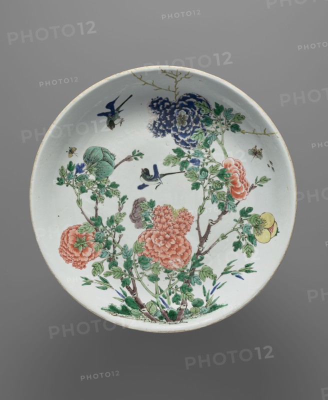Platter, signed. Decorated with images of birds and chrysanthemums. 19th century, China. Dish, diameter 35.5 cm., A round ceramic dish with a white background decorated with a botanical scene featuring a group of flowering branches with green leaves and stems, several large clustered blossoms in shades of coral and blue, a round yellow fruit, and a pale green bud; a few small blue and green birds are perched among the branches and flying, and a few small insects appear near the flowers; the rim shows a thin pale beige edge and the overall image includes colors white, green, coral, blue, yellow, beige, and brown.