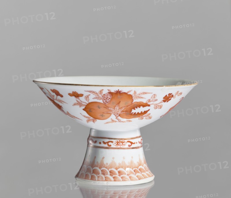 Platter, Chinese, 19th century. Porcelain platter on a tall, widening foot with white glaze, height 11.7 cm, diameter 19.5 cm. Decoration painted in red overglaze iron: central peach, pomegranate, and Buddha's hand in a centripetal composition; exterior shows three flowered branches bearing peach, pomegranate, and Buddha's hand motifs, a sign of happiness surrounded by four bats; ornamental bands on the foot. Colored porcelain, floral decorations, pomegranates, peaches, bats, happiness symbol., A white porcelain stem cup with orange decorative painting, showing a wide shallow bowl perched on a flared pedestal stem; the bowl rim has a thin gold edge and the exterior of the bowl is painted with orange floral and fruit motifs and leafy branches, while the pedestal carries orange ornamental bands and a repeating scallop-like pattern; the background is plain light gray.