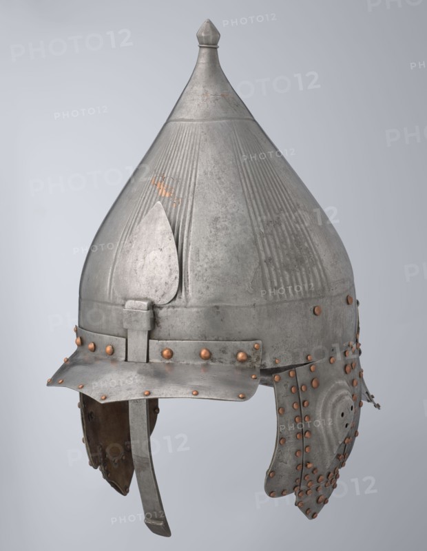 Helmet, 16th century, Turkey; Islamic, Turkish, Ottoman dynasty 1299–1922. Militaria headgear with movable cheekpieces and a nosepiece passing through the browband. Fluted bell. Browband and cheekpieces connected with copper rivets; neckpiece additionally with brass hinges. Weight 1.92 kg; height 39.0 cm; width 37.0 cm; depth 24.0 cm., A metal conical helmet with a pointed finial at the top, showing a weathered silver-gray surface with areas of darker gray patina and small patches of copper-colored wear; the helmet has vertical ridged decorations on the dome and a teardrop-shaped raised plate on the front, a brim that angles down slightly at the front, and a chin strap hanging below; a band around the lower dome is secured with a group of round copper-colored rivets, and additional copper-colored rivets decorate the ear guards and lower edges, which are segmented and show inner brownish leather lining.