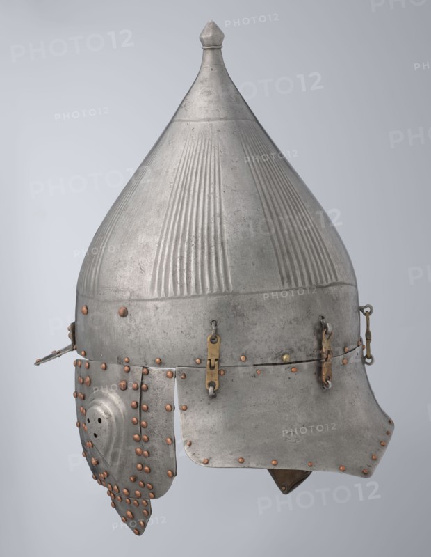 Helmet, Islamic art, Turkish, Ottoman dynasty 1299–1922, militaria headgear. Movable cheekpieces and a nosepiece passing through the browband. Fluted bell. Browband and cheekpieces connected with copper rivets; neckpiece additionally with brass hinges. 16th century, Turkey. Weight 1.92 kg; height 39.0 cm; width 37.0 cm; depth 24.0 cm., A metal helmet with a pointed conical top and a rounded dome featuring vertical ridged textures, mostly silver-gray in color with areas of darker gray patina, fitted with a lower brim and cheek guards. The surface shows numerous small rounded rivets in a coppery brown color arranged along edges and panels. There are a few small brass-colored metal fittings and hinges connecting parts of the helmet, including a few strap-like fasteners. One cheek guard has a curved shape with perforations and is attached by rows of coppery rivets. The overall object appears worn with slight surface scratches and variations in the silver-gray finish, set against a pale gra...