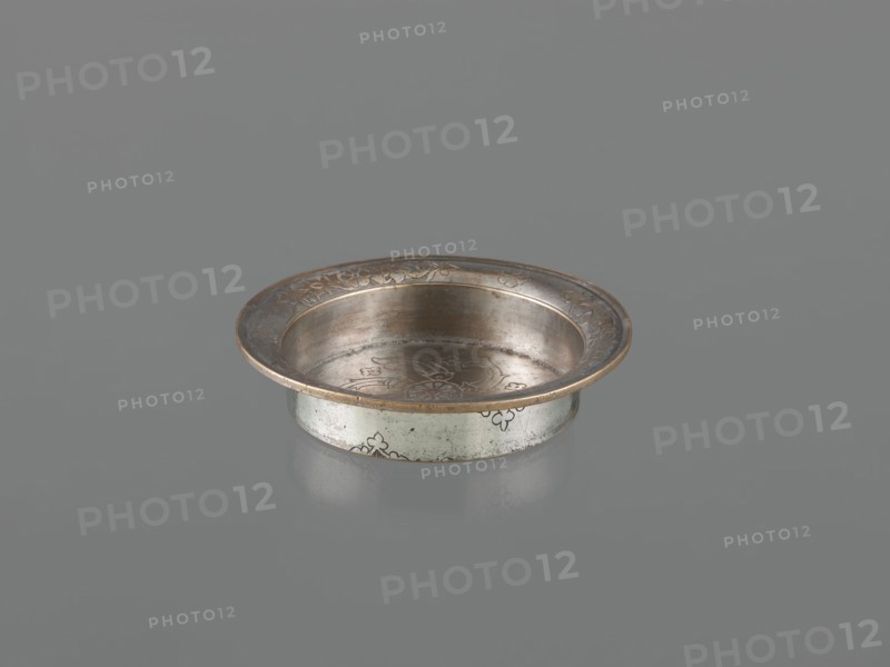 Bowl, Islamic art, metal bowl with fish, floral ornament and rosettes; flat bottom, low straight wall with slightly rounded rim; decorated with palmettes on the outer walls; 18th century; Turkey, / Iran ; vessel; height 4.4 cm, diameter 21.2 cm, A shallow round metal dish with a wide flared rim sits on a smooth gray surface, the dish showing a metallic palette of silver and warm bronze tones with subtle darker patina. The interior and rim display engraved floral and scroll motifs, and the exterior side bears small decorative patterns. The dish casts a soft reflection on the surface beneath it, and the overall scene contains shades of gray in the background and surface.