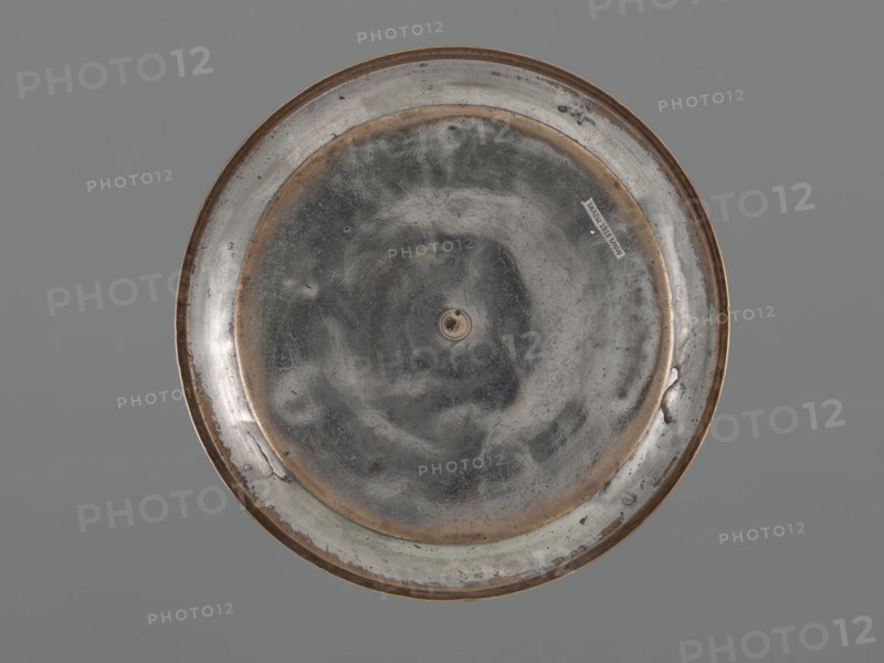 Bowl, Bowl with a flat bottom, low straight wall and slightly rounded rim; decorated with palmettes on the outer walls, fish, floral ornament, and rosettes. Metal. 18th century, possibly Turkey or Iran. Vessel; height 4.4 cm, diameter 21.2 cm., A round metal object viewed from above, centered against a flat gray background, with a worn reflective surface showing a mixture of silvery gray and darker gray tones and areas of brownish tarnish along the rim and inner ring; the surface has visible scratches and scuffs, a small central raised element, and a few irregular dark spots and blemishes around the edge.