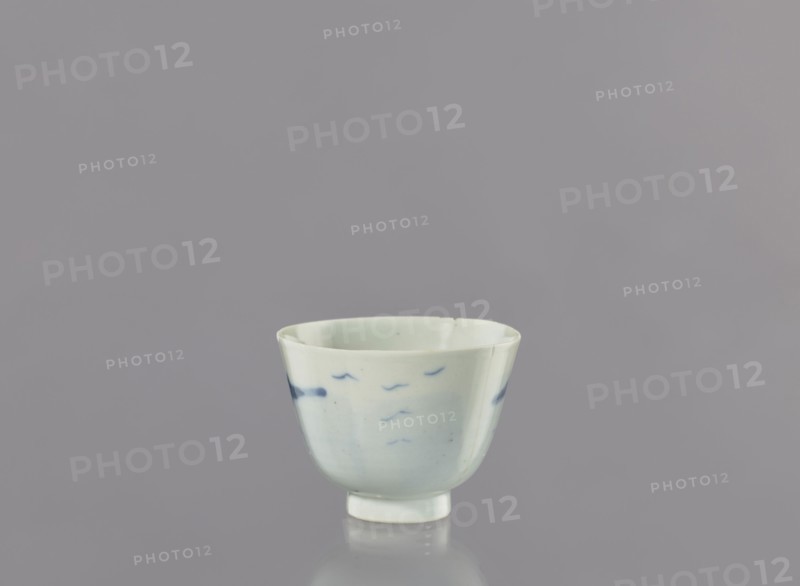 Bowl, Japanese, landscape motifs, ceramics, ca. 1750–1800, Japan, vessel; height 6.3 cm, diameter 8.3 cm, A small ceramic cup sits centered against a smooth gray background, the cup showing a pale off-white glaze with subtle blue brushstroke markings around its exterior, a few darker blue spots near the rim, a rounded body that tapers to a short foot, and a slight reflection visible on the surface beneath it; visible colors include gray, off-white, pale blue, and darker blue.