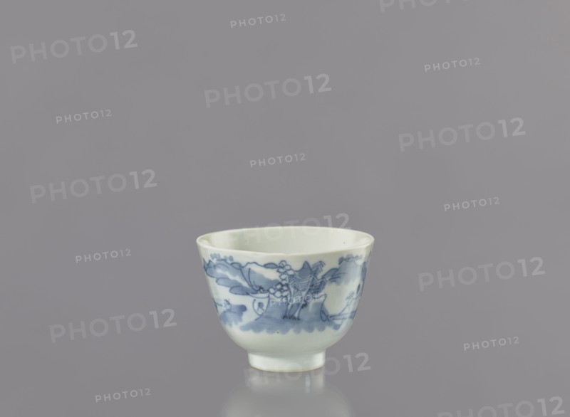 Bowl, Japanese porcelain bowl, widening toward the top, on a 0.5 cm foot. Decorated with a seaside landscape of fishermen among hills and coastal trees; two human figures—one with a fishing rod and the other with a net. Signature, on the bottom. Ca. 1750–1800, Arita, Japan. Vessel: height 6.3 cm, diameter 8.3 cm., A small porcelain cup sits centered against a uniform gray background, showing a pale off-white body with blue painted decorative motifs around its exterior; the cup has a rounded shape that tapers to a short foot, and the blue decoration includes flowing brushstrokes and floral or organic shapes in varying blue tones, with a smooth glossy surface that reflects light and casts a subtle reflection below.