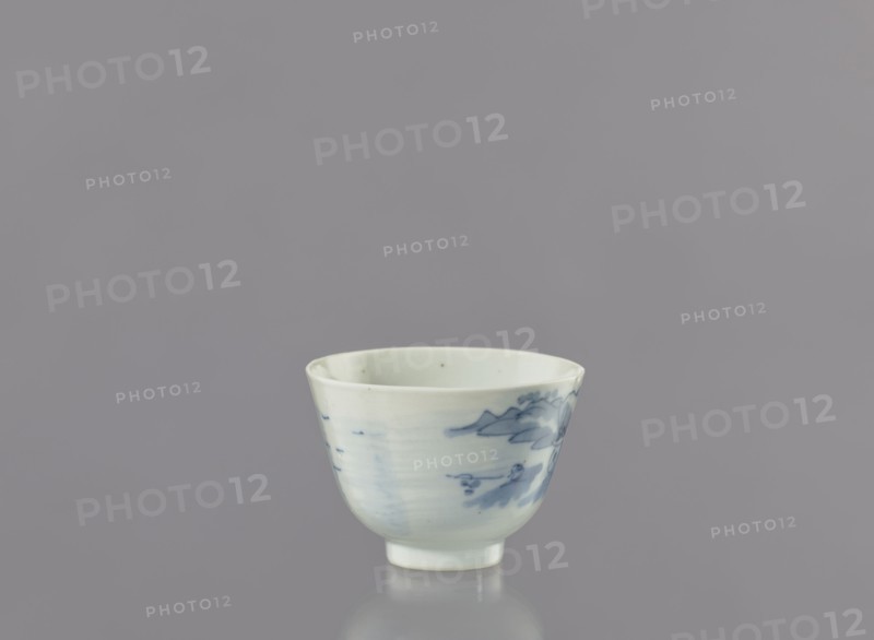 Bowl, Japanese, fishermen, seaside landscapes, bowls, landscape motifs, ceramics, unknown provenance. Porcelain bowl widening towards the top, on a 0.5 cm foot. Decorated with a view of fishermen on the seashore among hills and coastal trees; two human figures: one with a fishing rod and the other with a net. Signature, on the bottom. Ca. 1750–1800, Arita, Japan. Vessel height 6.3 cm, diameter 8.3 cm., A small ceramic cup sits centered against a smooth gray background, showing a pale off-white body with delicate blue-painted motifs on its exterior; the cup has a slightly flared rim, a rounded bowl shape narrowing to a short foot, and faint reflections on the surface beneath it.
