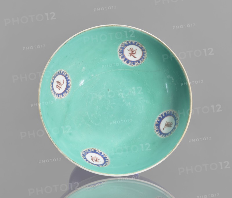 A footed porcelain bowl, Chinese, 19th century Qing Dynasty. Cobalt blue exterior glaze, green interior glaze; overglaze iron hieroglyphs, overglaze enamel paints, gilded edge and medal-gilding. Decorative vessel, height 12 cm, diameter 16.9 cm., and provenance. Categories: footed bowls, ceramics, colored porcelain, glazed ceramics., A shallow round dish with a smooth turquoise interior and a thin off-white rim, decorated with a few round medallions placed near the edge that have blue scalloped borders and white centers, resting on a reflective gray surface with a soft gray background; visible colors include turquoise, blue, white, off-white, and gray.