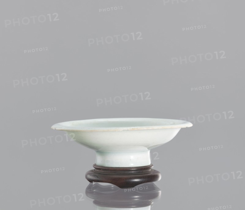 Alcohol bowl with base, Chinese, ceramics, porcelain vodka bowl. Plate with widely flared walls and a protruding rim. The bowl rests on a tall stem. Transparent glaze in a bluish tone. Flat, round base with a side cut at the bottom into three flat legs, made of profiled wood. 19th century, China. Vessel. Height 3 cm, diameter 10.3 cm., A shallow bowl with a wide rim and a short pedestal base sits on a carved stand; the bowl is a pale bluish-white color with subtle greenish tints and a thin slightly darker edge, while the stand is a dark brown wood; the background is a uniform light gray and the surface below the stand shows a soft reflection.