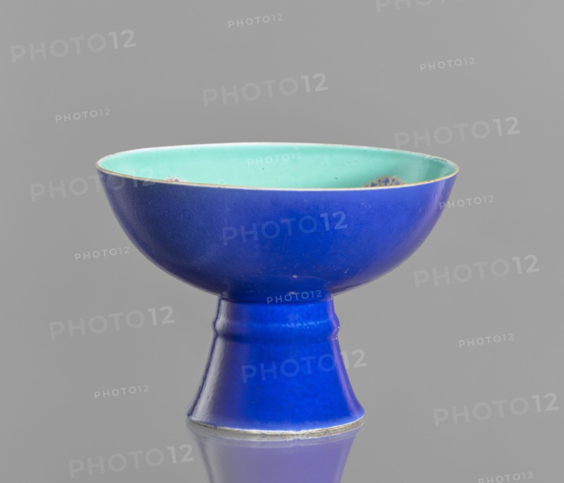 A tall footed porcelain bowl, Chinese, 19th century Qing Dynasty. Outer glaze stained cobalt, inner glaze green. Overglaze iron hieroglyphs, gilding, overglaze enamel paints, gilded edge. Height 11.8 cm, diameter 16.8 cm., and provenance., A shallow bowl with a flared foot, shown against a plain gray background, featuring a deep cobalt blue exterior and foot and a pale turquoise interior; the rim has a thin metallic edge and there are a few small blemishes and speckles on the interior surface, while the reflective surface beneath the object shows a muted blue reflection.