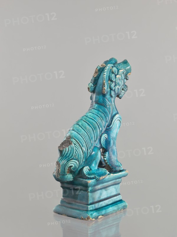 Lion fo, Chinese, Qing 1644–1911, 17th century, ceramic sculpture/figurine of a lion fo, sea green glaze, height 28 cm, animal figurine, iconography: fo., A turquoise-blue ceramic figurine of a seated mythical creature on a square pedestal, shown from the rear and slightly in profile; the creature has a mane of stylized curls and ridged, flowing fur along its back and tail, with visible worn areas where the glaze is chipped to reveal a darker underlying material; the pedestal and base share the same turquoise-blue color with lighter and darker variations, and the scene sits against a pale gray background with a subtle reflection on the surface beneath the pedestal.