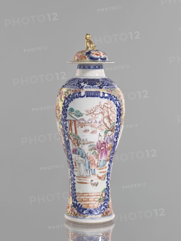 Vase with a lid, Chinese, ceramics, colored porcelain, 18th century. Height 28 cm. Lid topped with a modeled seated lion. Decoration: geometric and plant ornament around the neck; two large reserves bordered with plant ornament filled with figural genre scenes in a landscape, two smaller reserves with figures in a landscape; background of twigs with flowers on a gold ornamental ground. White glaze, underglaze cobalt, overglaze enamels in sepia, black, iron red, and gold. Vessel. Provenance unknown., A tall porcelain vase with a lid sits against a plain gray background, the body showing a glossy white surface decorated with intricate painted scenes framed by ornate blue floral borders; the painted imagery includes a group of people in traditional dress near a landscape with trees, water, and boats, rendered in muted colors such as blue, pink, green, brown, and gold accents; the lid is topped with a small sculpted figure finished in gold, and the rim and base also show decorative blue an...