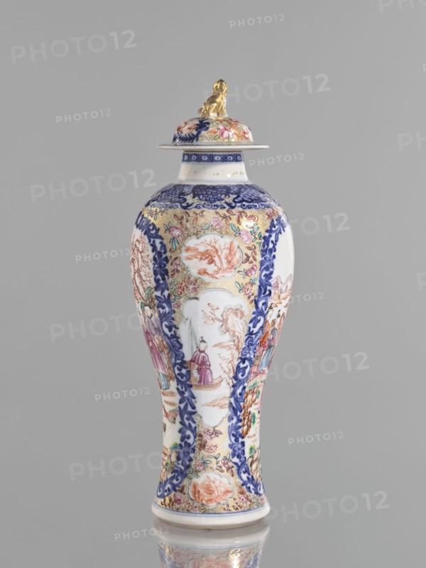 Vase with a lid, Chinese, 18th century. Porcelain vessel height 28 cm with white glaze, underglaze cobalt painting and overglaze enamels in sepia, black, iron red, and gold. Lid topped by a modeled seated lion. Decoration: geometric and plant ornament around the neck; two large reserves bordered with plant ornament containing figural scenes in a landscape, two smaller reserves with figures in a landscape; background of twigs with flowers on a gold ornamental ground. Genre scenes, floral decorations, and lions. Provenance unknown., A tall vase with a lid stands against a plain gray background, featuring ornate painted decoration in colors including white, blue, gold, pink, green, purple, and orange, with a small gold sculpted figure perched on the lid; the vase body tapers from a rounded shoulder into a narrow waist and flares slightly at the base, showing panels of floral patterns, scrolling blue borders, and several painted scenes with human figures and landscape motifs, and the lid a...
