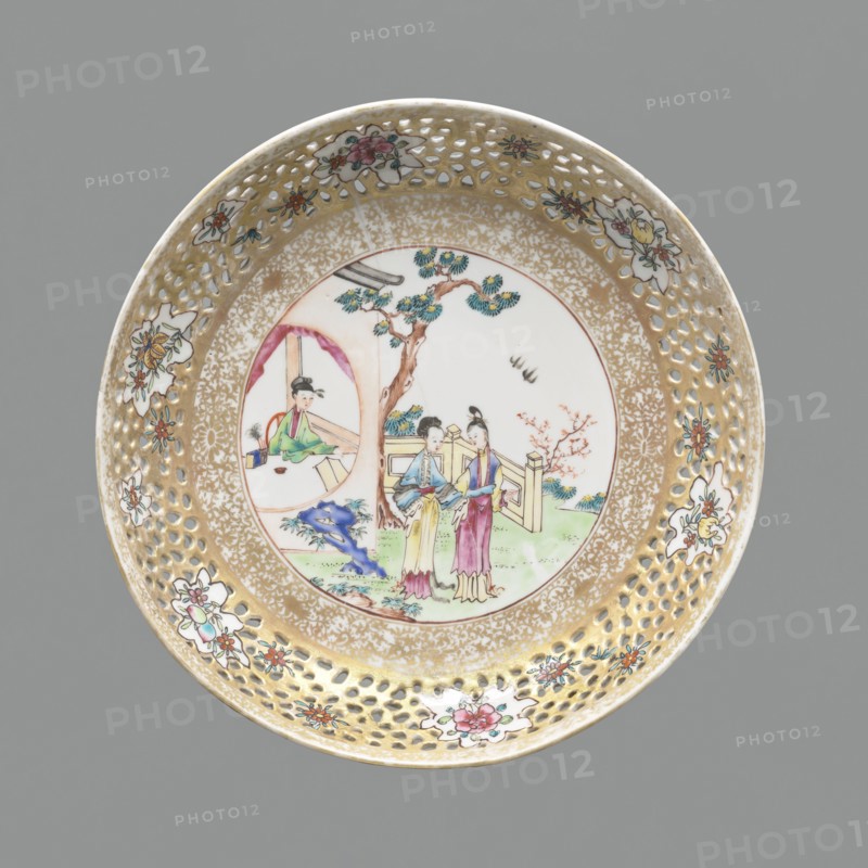 Plate, Chinese, figural scenes, openwork, colored porcelain, ceramics, plant ornaments, Qing 1644–1911. Openwork rim with plant projections flowers, fruits. White glaze. 18th–19th century, China. Vessel; height 3.5 cm, diameter 20.8 cm., A round decorative ceramic plate with a perforated rim and a painted central scene; the rim has a gold background with a pattern of small cutout holes and several floral motifs in white, pink, red, green, and blue, and the inner border shows a speckled gold design. The central round panel displays a courtyard scene with a tree in brown and green, a few figures wearing garments in pastel colors including yellow, pink, blue, green, and purple, and a small building element with a circular opening painted in muted tones; there is also a blue rock-like form, some grassy areas in green, and a small flowering branch in pink. The overall background behind the plate is gray.