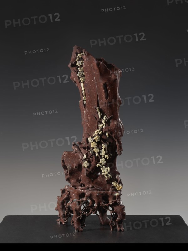 Vase on base, Chinese art, Chinese culture, Qing 1644–1911, huaniao style, plum trees, birds and flowers, brown soapstone, late 19th century, China, vessel, height 43.0 cm, A tall sculptural object with an irregular, porous form stands on a flat dark surface against a muted gray background; the sculpture is predominantly deep reddish-brown with a rough, pitted texture and flowing, carved cavities, and a winding cluster of small pale cream-colored blossoms and rounded buds ascends along its front, contrasting with the darker material.
