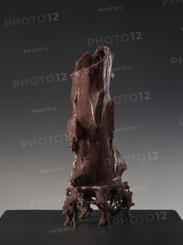 Vase on base, Chinese, Qing 1644–1911, huaniao style; decorated with plum trees, birds and flowers; brown soapstone; late 19th century, China; vessel; height 43.0 cm, A tall, irregularly shaped object with a glossy, deep reddish-brown surface standing on an openwork base of similar color; the object has a rough, carved appearance with smooth, flowing contours and several cavities and protrusions, with visible pale streaks and small pale flecks running across its surface; the base is organic and root-like with multiple arching supports and hollowed openings; the background is a smooth gradient ranging from dark charcoal at the top to a lighter gray toward the bottom, and the platform beneath the object is a flat black surface.