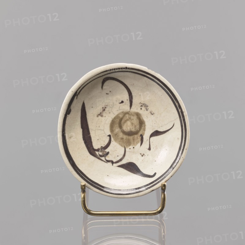 Plate, Southeast Asia culture, floral decorations, ceramics, trace of a Chinese inscription on the bottom, 18th–19th century, Southeast Asia place of origin, vessel, height 2 cm, diameter 10.5 cm, A shallow round ceramic dish with a cream-colored surface and a glossy finish sits on a small brass stand; the interior shows a decorative motif in dark brown and muted olive tones forming a central circular shape and flowing, curved brushstrokes that suggest organic, leaf-like forms, with a dark brown double border near the rim; the glaze shows fine crackling and a few small dark spots, and the background surrounding the dish is a uniform light gray.