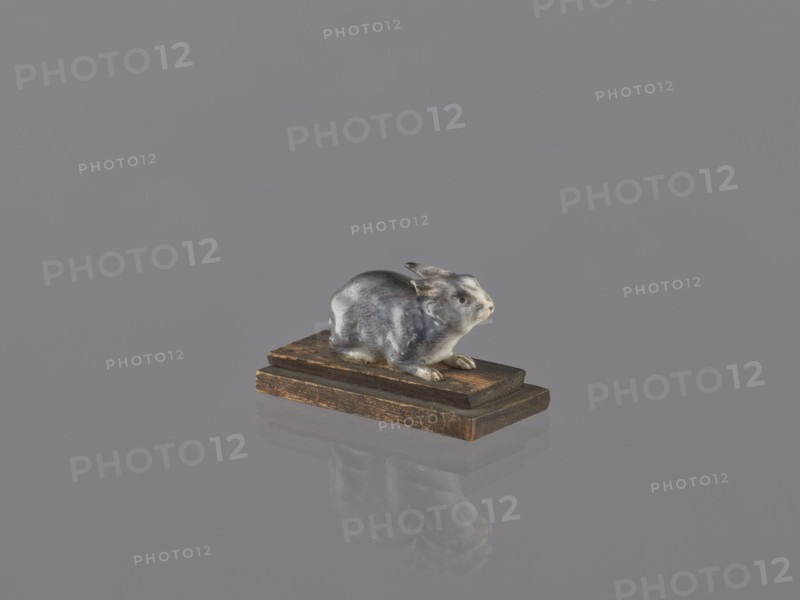 Rabbit, ceramic figurine/sculpture of an animal rabbit, ceramicist, Japanese, Meiji period 1868–1912, early 20th century, height 2.2 cm, width 4.1 cm; inscriptions/repossessions/, A small sculpted animal figure in shades of gray and white sits on a group of wooden blocks in brown tones; the figure has a glossy surface and a subtle reflection on the smooth gray background, with visible contours of ears, face, and paws, and the wooden base shows darker and lighter brown grain and worn edges.
