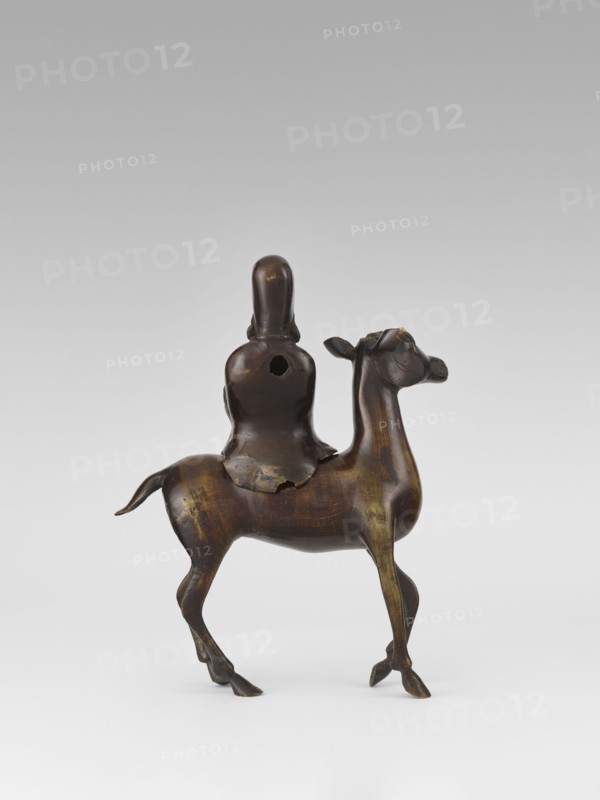 Shoulao on a deer — incense burner; creator, sculptor; Narozno Eckersdorf provenance; deer; Chinese art; Chinese culture; Taoism; incense burner; Shouxing iconography; sages; Qing dynasty 1644–1911, 18th century; China; sculpture; height 23.7 cm, A small sculptural object showing a rider seated on an animal that resembles a camel; the piece appears to be made of a dark brown material with areas of lighter brown and subtle yellowish highlights, and the surface shows a smooth, slightly glossy patina. The rider is simplified and rounded with a hooded or peaked head and a cloak draped over the shoulders, and there is a round opening visible on the rider's back. The animal has a slender neck, an elongated head with small ears, a curved tail, and thin legs with defined hooves, and the overall form shows gentle contours and polished wear on raised areas. The background is neutral and gradient, shifting from pale gray at the top to near white at the bottom.