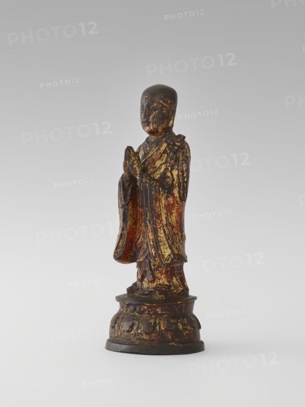 Monk, Chinese, Luohan/arhat figure, Buddhist art; monk/monastic subject on lotus throne; Qing dynasty 1644–1911, 19th century; sculpture/figurine, height 24.7 cm., A standing carved figure on a rounded base, with folded hands held near the chest, wearing flowing drapery; the surface shows worn gilding and traces of red and dark brown pigments, with areas of blackened patina, giving a mottled appearance of gold, red, dark brown, and black across the figure and base.