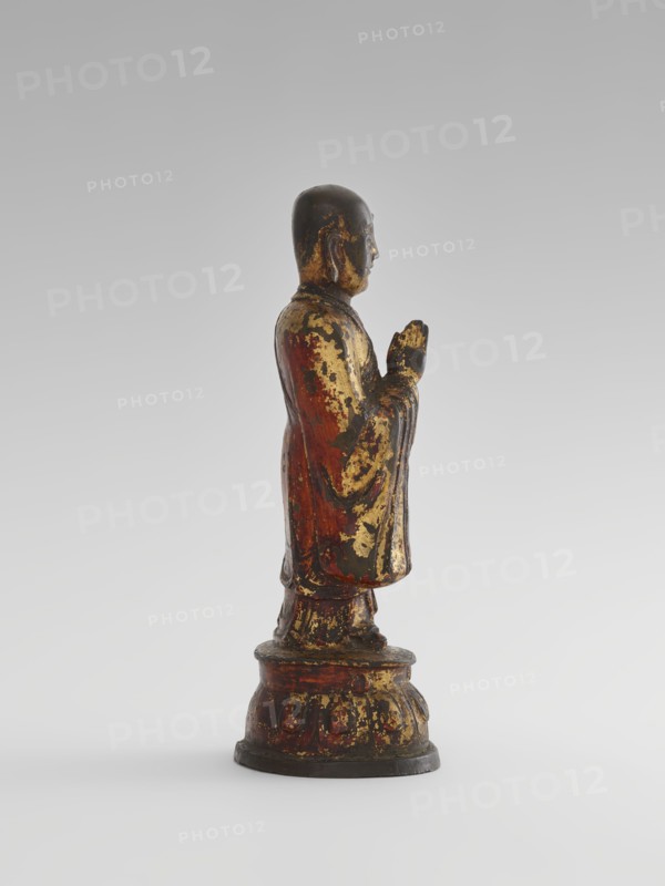 Monk, Chinese, Chinese art, sages, Luohan iconography, monks, arhat iconography, Buddhist art, lotus throne iconography, Qing 1644–1911, 19th century, China, sculpture, figurine, height 24.7 cm, A standing figurine shown in profile on a rounded pedestal, with a smooth rounded head and folded hands held near the chest; the surface shows worn patches of gold leaf over a base of reddish-brown and dark brown, with areas of black and muted green where the finish has aged; the robe has flowing vertical folds and the pedestal has carved rounded motifs with similar gold and reddish-brown wear; the background is a smooth gradient of light gray to off-white.