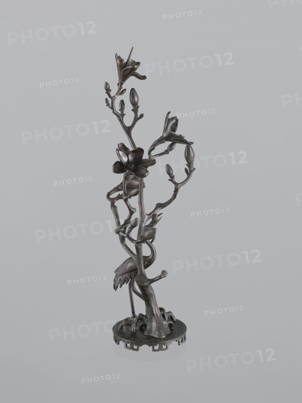 Candlestick shokudai, Japanese, magnolias, Meiji 1868–1912, candlesticks, plant tendril, cranes iconography, 2nd half of the 19th century, Japan, candlestick, height 49.5 cm, A dark metallic sculpture of a slender tree-like stem rising from a round base, with a group of branching twigs and buds and several open blossoms, a bird with a long neck and folded wings standing among the lower branches, and visible textures of bark and feather; the colors visible are dark brown and black against a light gray background.
