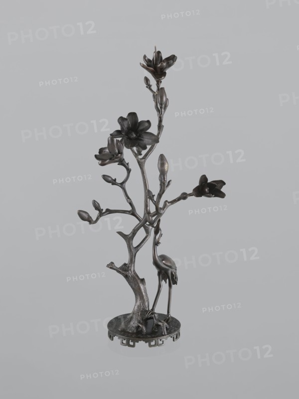 Candlestick shokudai, Japanese, magnolias, Meiji 1868–1912, candlestick with plant tendril and cranes iconography, 2nd half of the 19th century, Japan, height 49.5 cm, A metal sculpture depicting a slender, branching tree with a textured trunk rising from a round base, featuring several stylized blossoms and buds on its branches and a long-legged bird standing beside the trunk; the piece appears in dark bronze tones with hints of metallic sheen against a light gray background.