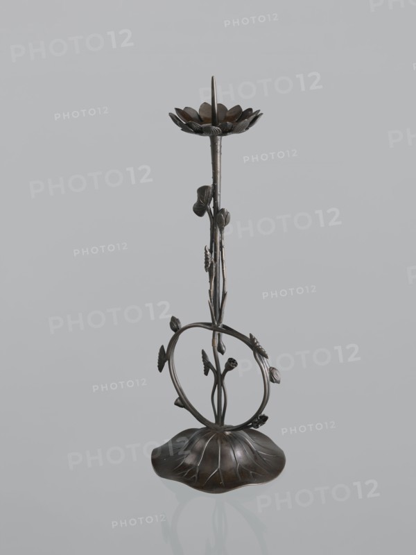 Candlestick shokudai, Takimoto, Iwami; Japanese art; Japanese culture; lotuses, leaves; candlestick; Meiji 1868–1912, 2nd half of the 19th century; Japan; unknown origin; height 44.0 cm, A sculptural metal object resembling a tall, slender stem rising from a rounded, petal-like base with a dark brown to bronze patina, featuring a looping circular vine element mid-height with several small leaf and bud shapes attached, a vertical stem with textured nodes and leaves, and a flower-like dish at the top with radiating petal forms and a pointed central spike; visible colors include dark brown, bronze, and subtle black accents set against a pale gray background.
