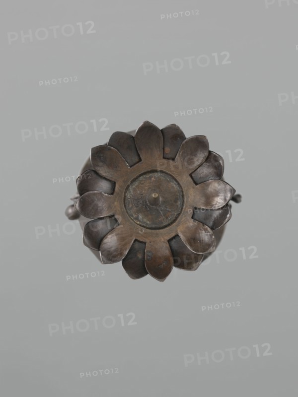 Candlestick shokudai, maker Takimoto, Iwami; Japanese; decoration: lotuses and leaves; Meiji period 1868–1912, 2nd half of the 19th century; Japan; material/role: candlestick; height 44.0 cm; provenance unknown., A close-up view of a round, flower-shaped metal object with petal-like segments radiating from a central circular disk; the metal shows a dark brown and bronze patina with areas of lighter brown and subtle greenish oxidation; the petals have smooth, slightly curved surfaces and varying reflections that suggest wear; the background is a uniform soft gray.