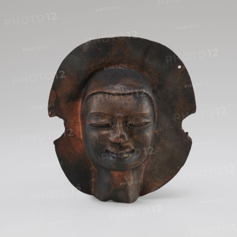 Head, unknown provenance; Tibetan culture; monks, disciples; Tibetan Buddhist art, Buddhist art; faces; 19th–20th century; Tibet place of origin?; figurine sculpture; height 8.9 cm, width 3.5 cm, A sculpted face mask with a smooth, rounded forehead and closed eyes set within a broad, irregular halo-like backplate, showing a serene, slightly smiling mouth and a rounded chin; the surface displays a mix of dark brown, rusty orange, and black patina with patches of lighter brown and subtle metallic sheen, and the backplate has irregular edges and a small perforation on one side with areas of wear and mottled coloration.