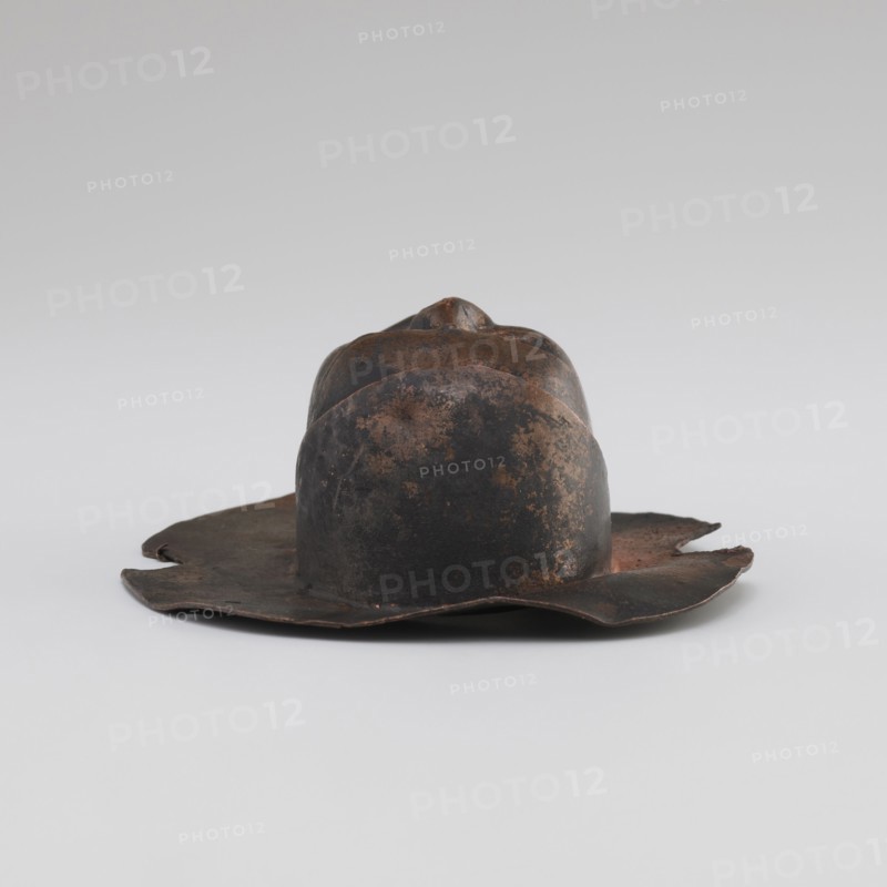 Head, unknown provenance; Tibetan; monks, disciples; Tibetan Buddhist art, Buddhist art; faces; 19th/20th century; Tibet place of origin?; figurine sculpture; height 8.9 cm, width 3.5 cm, A worn metal hat with an irregular brim and a domed crown sits on a neutral surface; the object shows a mottled surface with shades of dark brown, rusty orange, and black, with areas of uneven texture and small patches of lighter brown.