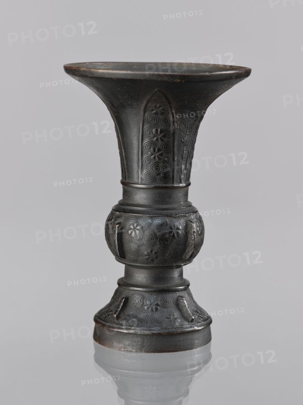 Gu-type vase, gu cups, Chinese art, Chinese culture, ceremonial and ritual vessel decorated with spiral and plant motifs, Qing dynasty 1644–1911, 18th century, China, vessel; height 14.0 cm, diameter 10.0 cm, A dark metal vase with a flared rim and a narrow waist sits centered against a pale gray background, showing a rounded bulbous midsection and a stepped base; the surface of the vase displays decorative embossed spiral and floral motifs and vertical recessed panels, with visible wear and slight brownish highlights on edges, and a faint reflection of the vase on the glossy surface beneath.