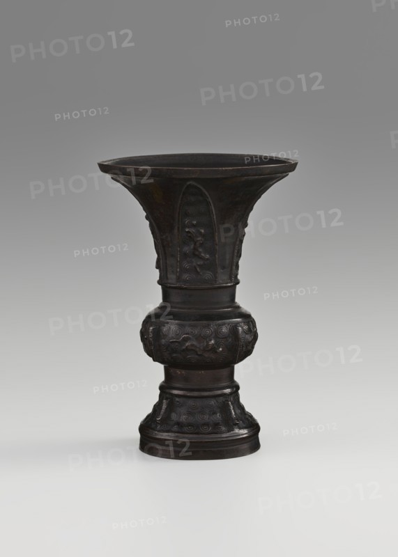 Gu-type vessel, gu cups, Chinese art, Chinese culture, ceremonial vessels, Qing 1644–1911, ritual vessels, 18th century, China, vessel; height 15 cm, diameter 10.7 cm, A dark brown metal vessel with a flared rim and a tall, tapering body standing on a rounded base, featuring raised decorative panels and carved motifs on its upper and lower sections, shown against a neutral gray background that fades from darker at the top to lighter toward the bottom.