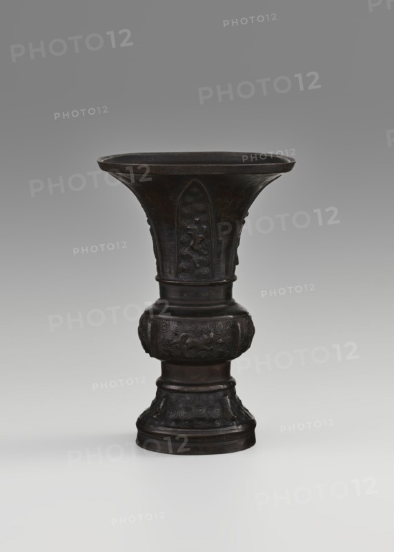 Gu-type vessel gu cup, Chinese, Qing dynasty 1644–1911, 18th century, ritual/ceremonial vessel, height 15 cm, diameter 10.7 cm., A dark bronze vessel with a wide flared rim tapering into a narrow waist and then expanding into a rounded midsection before narrowing again to a decorated base, standing against a neutral gray gradient background. The surface of the vessel shows intricate raised decorative motifs in repeated swirling patterns and stylized animal or mythical creature figures on the midsection and upper body, with carved vertical panels on the flared portion. The overall color palette is dark brown to nearly black with subtle lighter brown highlights on raised areas, and the background is shades of gray.