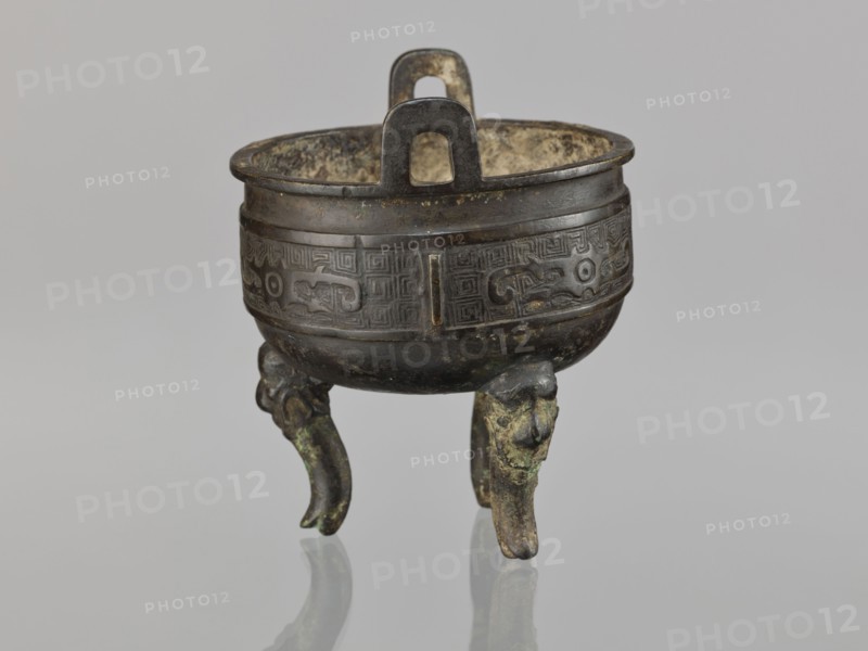Ding incense burner, Chinese, ding vessel, ceremonial/ritual vessel, decorated with leiwen ornament, meander ornament, and dragons; Qing dynasty 1644–1911, 19th century, China; vessel; height 12.8 cm, diameter 10.5 cm., A small bronze vessel stands on a reflective surface against a pale gray background, showing a rounded bowl with a wide rim and a pair of vertical loop handles rising above the rim; the body has a decorative band with incised patterns and raised motifs, and the vessel rests on three short, curved legs that resemble stylized animal forms. The visible colors are dark brown and black on the metal, with areas of greenish corrosion and lighter brown or tan patina on the interior and raised surfaces, and the background and reflection are shades of light gray.