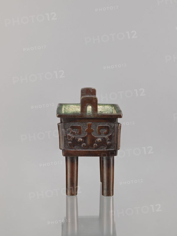 Fangding incense burner, Narozno Eckersdorf, Chinese, Qing dynasty 1644–1911, 18th century. Bronze ceremonial/ritual vessel fangding standing on four cylindrical legs, decorated with meander and leiwen ornament and taotie iconography. Dimensions: height 15.3 cm, width 13.1 cm, depth 10.3 cm., A small tripod vessel made of dark brown metal with a square body and a flat rim, standing on three cylindrical legs; the exterior of the body features a carved decorative pattern with raised rounded elements, and the interior has a greenish patina; a rectangular loop handle rises from the center of the rim; the background is a smooth pale gray.
