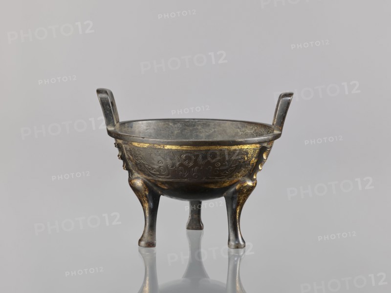 Ding incense burner, Chinese; decorated with fantasy beasts, meander and leiwen ornament; Qing dynasty 1644–1911, 19th century, ceremonial/ritual vessel censer; height 12.6 cm, width 16 cm, diameter 14.5 cm., An aged metallic vessel with a round bowl supported by three curved legs and two upright handles, showing a dark gray and bronze patina with areas of gold-colored accents and worn highlights; the bowl's exterior surface has subtle raised decorative patterns and worn gilding, and the interior appears darker and more matte; the object is photographed against a smooth light gray background with a faint reflective surface below producing a soft mirror image.