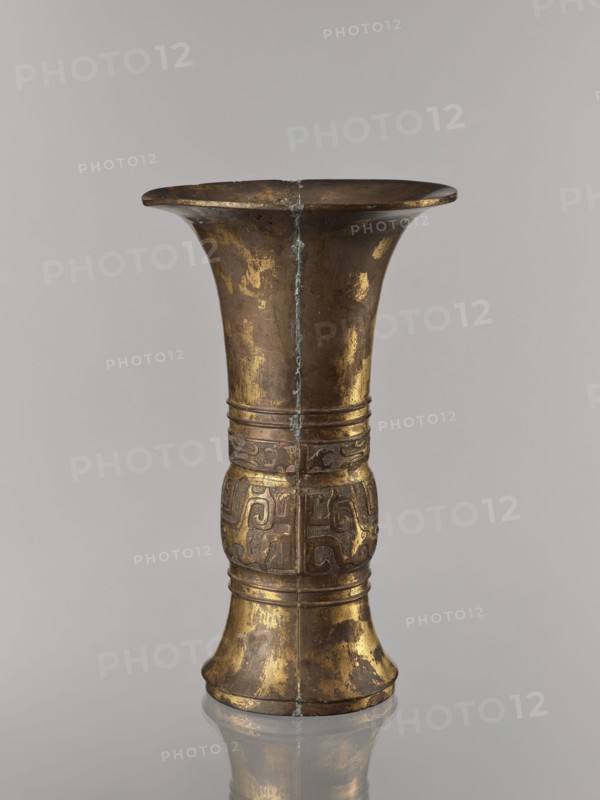 Zun vase; maker, Chinese; ceremonial vessel vessel, cup; taotie and fantastic beasts iconography; meander and leiwen ornament; Qing dynasty 1644–1911, 19th century ; China; height 31.7 cm, diameter 19.0 cm., A tall metallic vessel with a flared rim and a narrower waist, showing worn patches of gold and brown tones across its surface; a vertical seam runs down its center and there are decorative raised bands and a band of carved or embossed scroll-like patterns around the midsection; the metal shows areas of tarnish and patina in darker brown and greenish hues, and the piece sits on a reflective surface that mirrors its base; the visible colors include gold, brown, dark brown, greenish, and gray.
