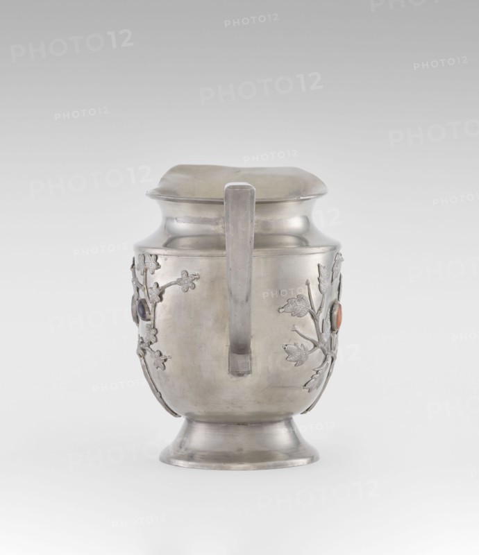 Jug, plant ornament, Chinese, Chinese art, teapot form, fruit ornament, applied plastic decoration: flowering, leafy bunches with oval set semi-precious stones, plum trees motif, Qing dynasty 1644–1911, 19th century, China; vessel, height 20.6 cm, width 23.0 cm., A metallic pitcher with a smooth, silvery surface sits against a pale gray background; it has a flared base, a rounded body, a slightly flared neck, and a square-edged handle running vertically down the front. A few raised floral and leaf decorations are attached to the sides of the body, and small round cabochon-like accents in dark and orange hues are set within the floral motifs. The overall colors visible are silver, gray, dark bluish, and orange.