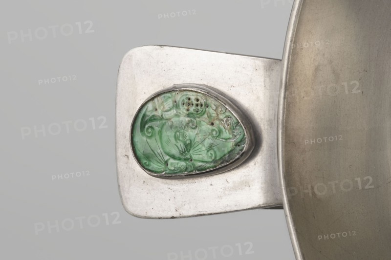 Vessel, Chinese, plant ornament and decorative motifs including Chinese symbols, Qing dynasty 1644–1911, 19th century, China; vessel; height 8.3 cm, diameter 20.0 cm, A close-up of a metallic object showing a rectangular silver-toned surface with rounded corners and a raised oval setting holding a green, carved stone with swirling, organic patterns, paired with a curved silver rim of a larger vessel visible at the right edge; colors present include silver, green, pale gray, and hints of darker gray and brown.