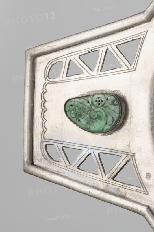 Platter, Chinese, openwork with bats and coins iconography, Qing dynasty 1644–1911, 19th century, China. Platter on a foot with two trapezoidal handles; vessel. Height 48.8 cm; width 24.0 cm., A close-up of a metallic object with a pale silver surface and a slightly tarnished finish, featuring cut-out triangular and rounded shapes and scalloped decorative engraving along one edge; near the center is a green, teardrop-shaped stone with carved swirling floral motifs set into a dark bezel, and the background is a smooth light gray.