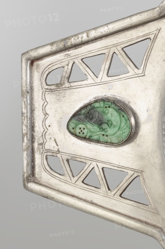 Platter, Chinese, openwork with bats and coins iconography, Qing dynasty 1644–1911, 19th century. Platter on a foot with two trapezoidal handles, vessel. Height 48.7 cm; width 24.2 cm., A close-up of a metallic object showing a section of its decorative surface in silver tones with areas of tarnish and darker discoloration; the design includes several triangular cutouts and scalloped engraved lines, with a green carved stone set into an oval bezel at the center that shows carved floral-like patterns and small perforations; the background is a smooth light gray.