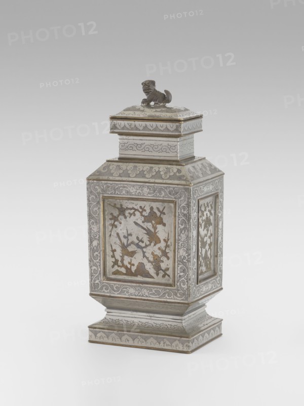 Teapot with a lid, Teapot with a profiled foot and neck, with a rectangular lid topped with a modeled lion figurine. Decorated with panels filled with a motif of a blossoming plum tree and birds, bordered with plant twigs panel motif engraved and set with metal. Foot, neck and lid decorated with plant ornament on a powdered background. Materials/style references: Chinese, huaniao style, Qing 1644–1911, 19th century. Iconography: lions, birds and flowers, fo, clouds, plant twig. Function: vessel teapot. Dimensions: height 27.3 cm, width 11.5 cm, depth 8.3 cm., A decorative metal container with a square, tapered body and a matching lid topped by a small sculpted animal figure, shown against a light gray background. The container has ornate engraved and inlaid panels on each side featuring floral and bird motifs, with borders of scrolling foliage and patterned bands. The lid and stepped base carry similar engraved patterns and flourishes. Visible colors include silver, gray, and accents o...