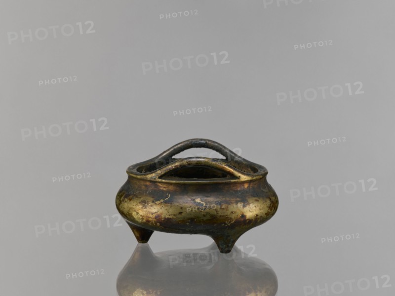 Li-type incense burner, Chinese, ceremonial Li-type ritual vessel, Ming style, Qing 1644–1911, backdated signatures, 19th century, China; vessel; height 6.8 cm, diameter 12.9 cm, A small metal vessel with a rounded body and a pair of curved handles across its open top sits on a reflective surface, showing a mirrored underside; the vessel has an aged, mottled finish with areas of gold, dark brown, and greenish patina, and it stands on three short tapered legs against a plain gray background.
