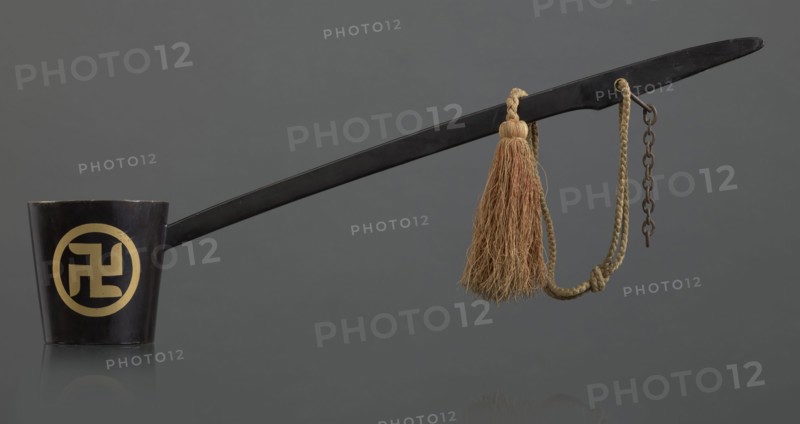 Ritual ladle, Chinese, Qing 1644–1911, Chinese Buddhism, ritual vessel with swastikas painted gold. A chain and silk cord with a tassel hang from the handle. Late 19th century, China. Length 57.5 cm., A long black ladle-like object extends across the image, attached at one end to a short black cup with a circular pale yellow decorative motif on its side, and at the other end to a braided tan cord that holds a tassel of coarse light brown fibers and a short rusty metal chain; the background is a uniform dark gray and the surface beneath the objects reflects them faintly.