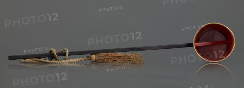 Ritual ladle, Chinese, Qing dynasty 1644–1911, late 19th century. Buddhism ritual vessel, painted gold swastikas; a chain and silk cord with a tassel hang from the handle. Length 57.5 cm., A long dark rod lies horizontally across a smooth gray surface with its reflection visible, attached near one end to a braided beige cord that forms a loop and leads to a tassel of coarse light brown fibers bound by a small beige cap; at the opposite end the rod is fitted into a shallow round cup that is red inside with a thin golden rim and a short red crossbar across its interior.