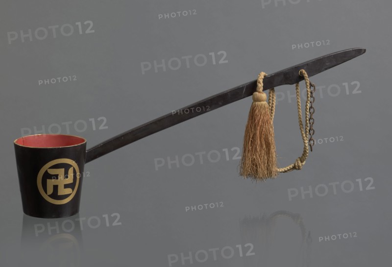 Ritual ladle; maker, Chinese; Qing dynasty 1644–1911; Chinese Buddhism ritual vessel; late 19th century, China; materials: chain and silk cord with a tassel hang from the handle; swastikas painted gold; length 57.5 cm., A long dark handle attached to a dark cup with a red interior rests against a gray background, with a gold-colored circular emblem on the cup's exterior partially visible; a braided tan cord with a tassel of natural brown fibers hangs near the handle's end alongside a short dark metal chain, and the scene reflects faintly on the surface below.