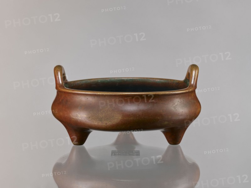 Li-type incense burner, Chinese, ceremonial vessel, Ming style Ming 1368–1644, Qing 1644–1911, Xuande 1426–1435, antidated signatures. Cast in golden-red bronze with light reddish-brown patina. Flattened low body with twig-shaped handles and low legs. Signed. 18th century, China. Vessel censer. Height 13 cm, diameter 26 cm, depth 6.5 cm., A low, rounded three-legged vessel with two upward-curving handles sits on a reflective surface; the vessel's exterior shows a brownish-bronze patina with areas of darker greenish and golden tones, and the interior appears dark with hints of green; the background is a smooth light gray.