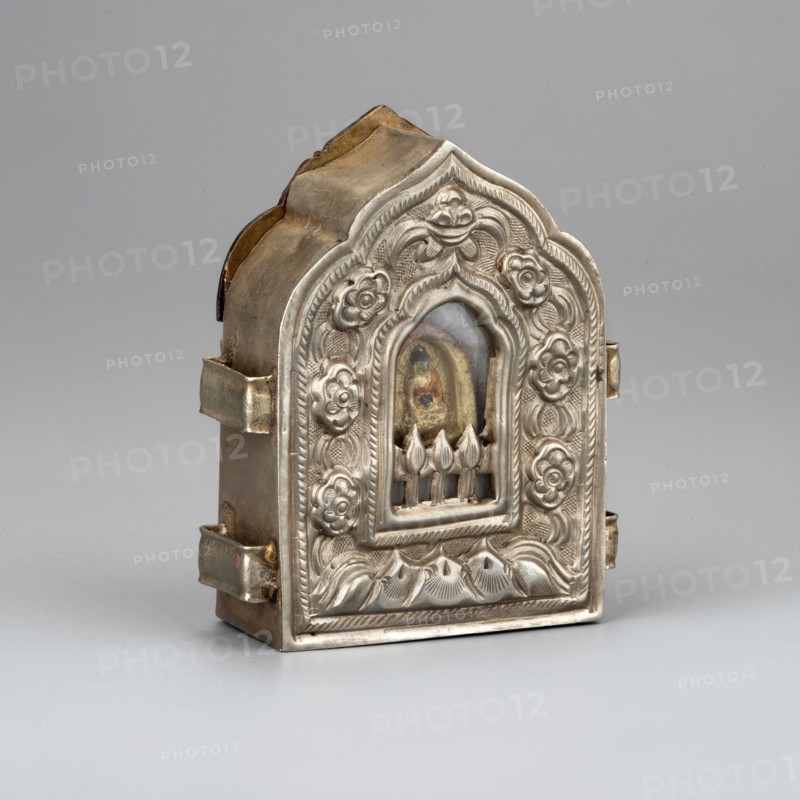 Ga'u box with a Buddha figure inside; maker, Himalayan Tibetan Buddhist art; amulet/cult object; Buddha iconography; floral ornament; inscriptions. Materials: thinly silvered, repoussé, chiseled and bonded copper sheet; glass pane; clay figurine originally probably silver-plated. Date: 19th century . Place of origin: Tibet . Dimensions: height 5.5 cm; width 4 cm., A small ornate metal box with a domed top and arched front sits against a neutral gray background; the box is primarily silver in color with areas of aged brass or gold tones, and features an embossed floral and leaf pattern surrounding a recessed arched window that reveals a small painted element inside with muted greens, reds, and brownish tones; the sides show metal straps and hinges with a slightly tarnished appearance, and the base rests on the flat surface casting a soft shadow.