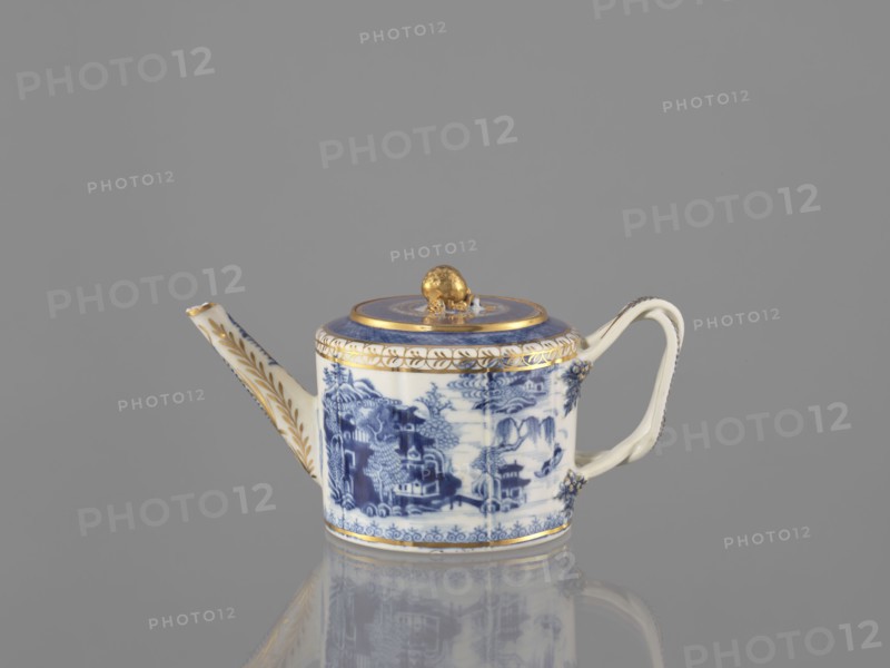 Teapot with a lid, Chinese, Qing dynasty 19th century, 1644–1912, blue-and-white porcelain teapot from a 22-piece tea and coffee service teapot, 6 cups, 6 bowls, 6 saucers, a bowl, a lid, and a plate, decorated with landscape motifs; ceramics, vessel; height 11 cm, width 21.9 cm, diameter 11.6 cm., A porcelain teapot sits against a neutral gray background, featuring a round body with a curved handle and a long tapered spout. The teapot displays blue and white painted scenes on its body and lid, with decorative gold trim around the rim, lid edge, and spout, and a gold knob on the lid. The handle and spout have gold accents and small floral-like applied details where they join the body. The overall colors visible are blue, white, gold, and gray from the background and reflective surface.