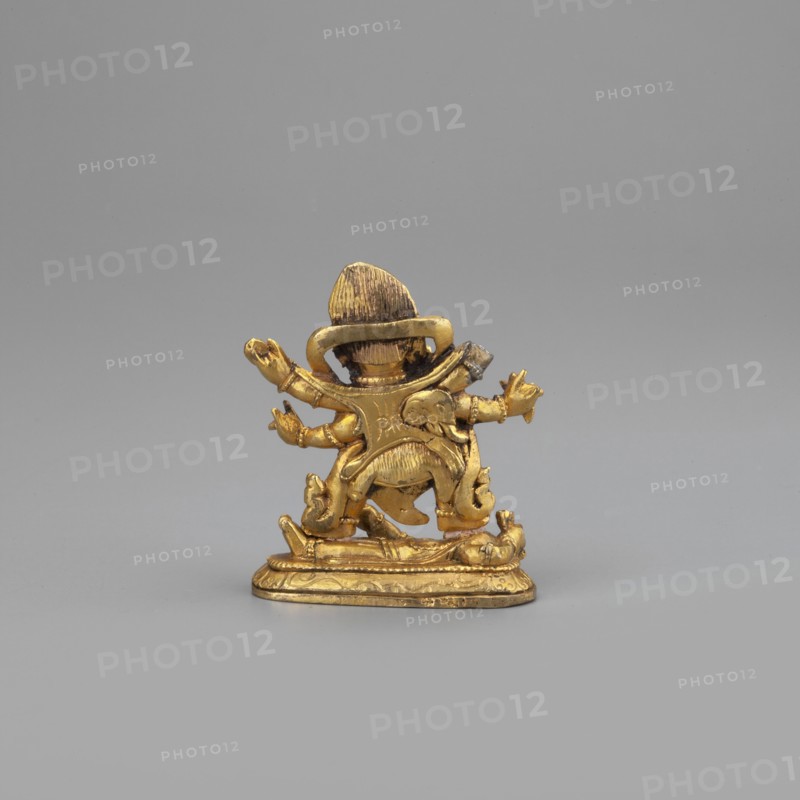 Six-armed Mahakala, unknown sculptor, 2nd half of the 19th century, Tibetan Buddhist Himalayan art; dharmapala iconography; Mongolia place of origin; sculpture, figurine; height 3.8 cm, width 3.1 cm, A small gold-colored metal figurine stands on a flat decorative base against a plain light gray background; the figurine shows a rounded head with a hat-like top, a curved collar or halo behind the head, a torso with sculpted folds and ornamentation, multiple outstretched arms with open hands, and stylized lower limbs and flowing drapery details. The visible colors are gold on the figurine and light gray in the background.